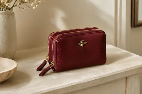 Porte-monnaie en cuir rouge vin à deux fermetures zippées, posé sur une console crème.