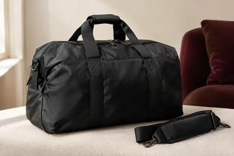 Sac de voyage noir surpiqué avec poignées et bandoulière amovible, posé dans un intérieur sobre aux murs crème.