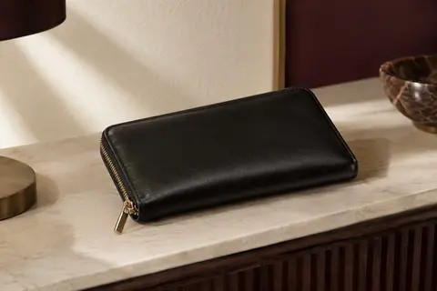 Portefeuille long noir en simili cuir posé dans un intérieur crème, avec fermeture éclair métallique visible.